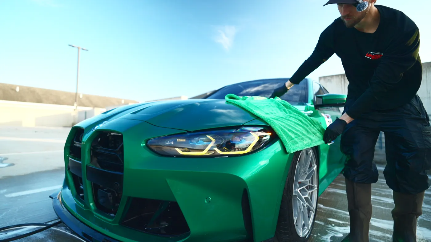 Detailer drying the headlight area of a green BMW with a green microfiber towel on a wet rooftop lot